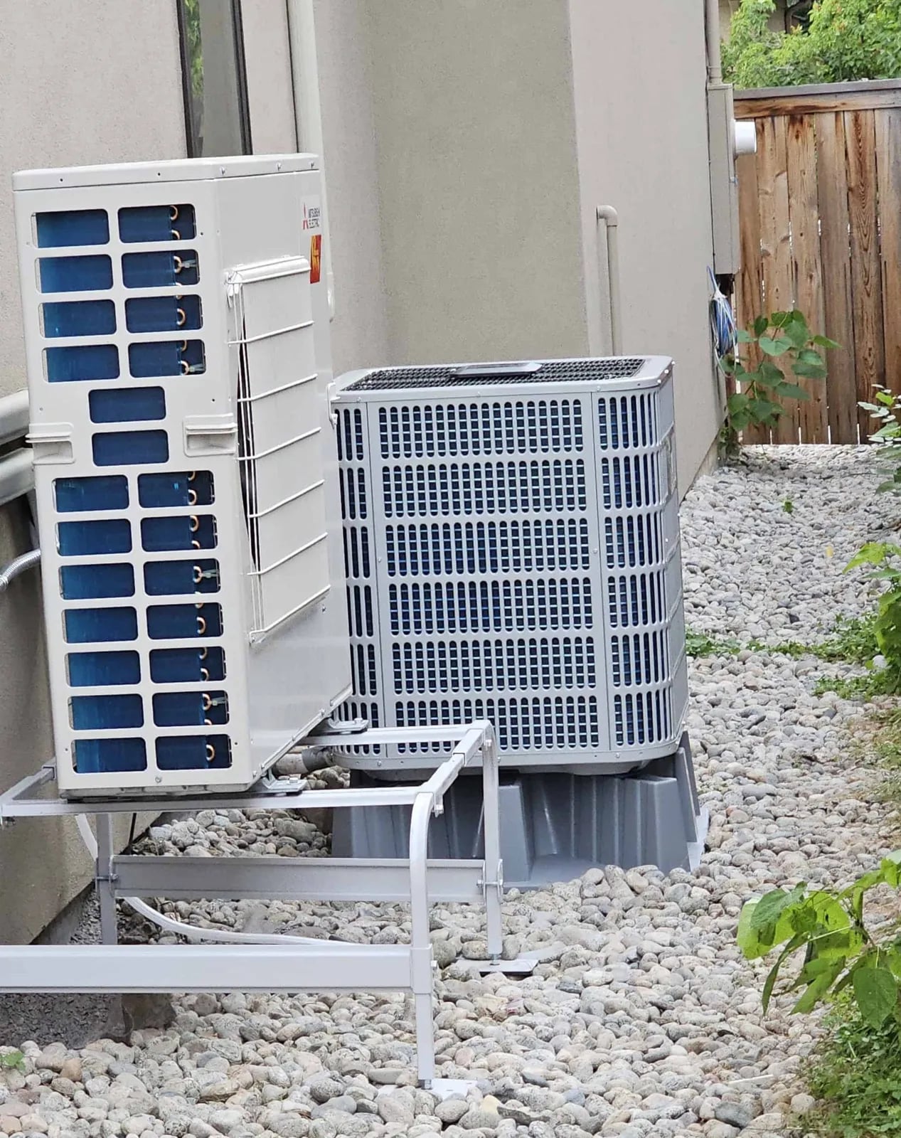 A Mitsubishi ductless outdoor unit on a wall stand next to a central heat pump on a riser pad over river rock beside stucco.